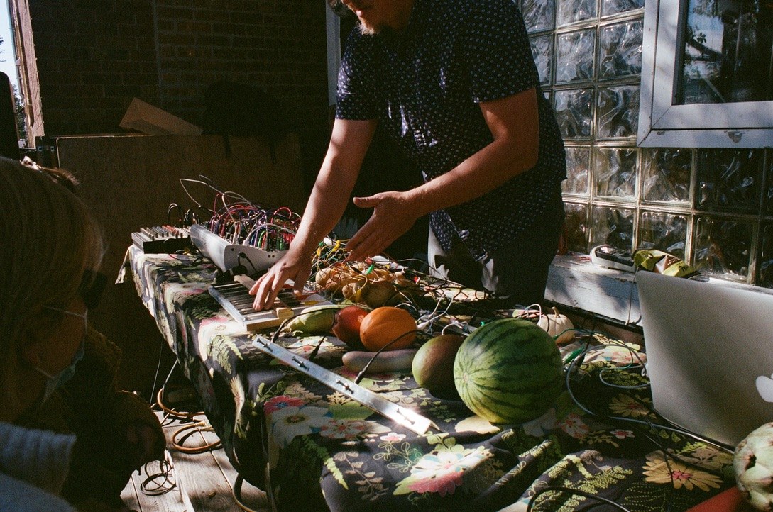 Hands playing a modular synthesizer wired into fruits and vegetables on a table